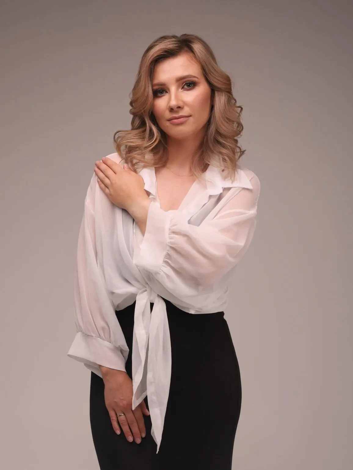 Une femme en blouse blanche légèrement transparente et pantalon noir pose sur un fond neutre.
