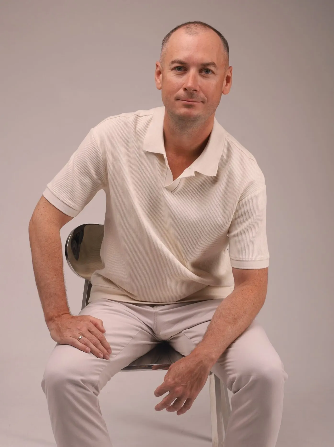 Un homme en tenue claire est assis sur une chaise devant un fond neutre.